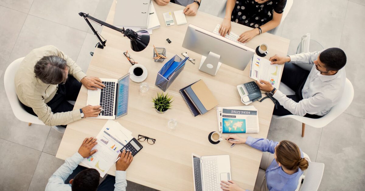 Overhead view of a small business team at a conference table using laptops and reviewing reports, planning software integration and productivity tools.